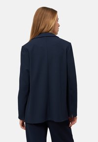 Blazer bleu marine à coupe ajustée, légèrement épaulé, avec une texture lisse. Doté d'un col cranté classique et de fentes latérales.