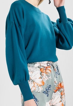 Personne portant un pull à manches longues bleu sarcelle avec des manches bouffantes et une jupe à grands motifs floraux orange et blanc.