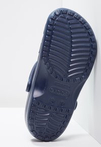 Semelle d'une chaussure Crocs bleu marine montrant un motif de bande de roulement texturé et des indications de taille sur fond blanc.