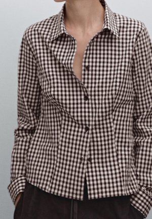 Personne portant une chemise à carreaux noire et blanche avec les boutons du haut déboutonnés et les mains dans les poches d'un pantalon foncé.