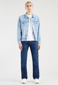 Jauns cilvēks ar īsiem blondiem matiem, ģērbies gaiši zilā džinsu jakā, baltā T-kreklā, tumši zilās džinsās un baltās kedās, stāvot pret baltu fona.