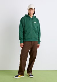 Green zip-up hoodie with front pockets, paired with brown pants and multicolored sneakers. Model wearing a beige cap, standing on green background.