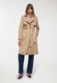 Trench coat beige con vita cintata, ampi revers e una texture morbida, abbinato a jeans a gamba larga blu scuro e mule marroni.