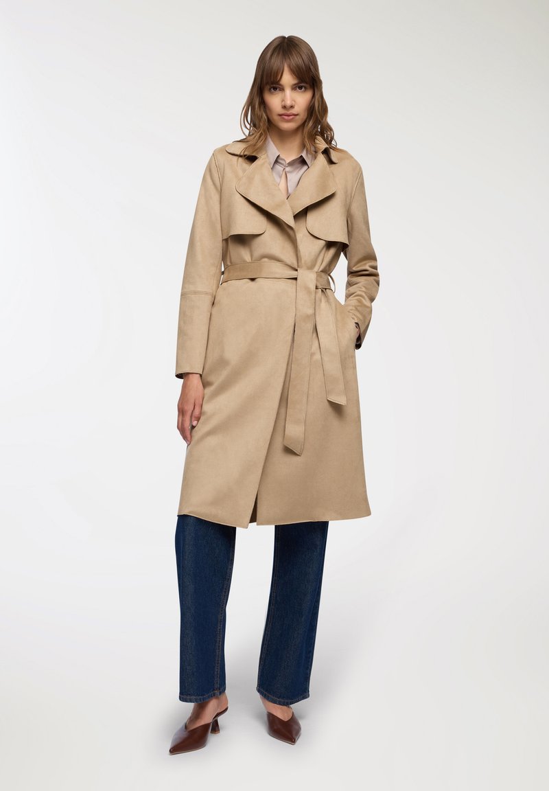 Trench coat beige con vita cintata, ampi revers e una texture morbida, abbinato a jeans a gamba larga blu scuro e mule marroni.