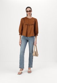 Blusa de algodón marrón con cuello volante, mangas abullonadas y detalles bordados, combinada con jeans rectos azul y bolso beige.