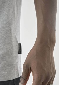 Graues Baumwoll-T-Shirt mit kurzen Ärmeln, ausgestattet mit einem seitlichen Etikett. Glatte Textur, runder Ausschnitt und lässige Passform. Getragen mit schwarzen Hosen.