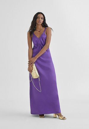Vestido maxi de satén púrpura con un frente retorcido, tirantes finos y una textura suave. Acompañado de un pequeño clutch metálico dorado y tacones.