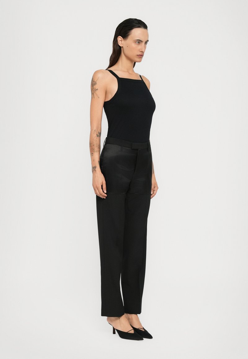 Femme aux longs cheveux noirs et aux tatouages, portant un haut sans manches noir, un pantalon taille haute noir et des escarpins noirs pointus, se tenant de côté.