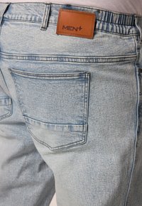 Jeans de mezclilla de color azul claro con un parche de cuero etiquetado "MEN+" en la cinturilla. Cuenta con dos bolsillos traseros con costuras visibles.