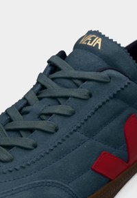 Κοντινό πλάνο μπλε βελουτέ αθλητικού παπουτσιού Veja με κόκκινο λογότυπο "V", μπλε κορδόνια που ταιριάζουν και χρυσό όνομα της μάρκας στη γλώσσα.