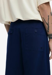 Pantaloni blu navy in cotone con una texture liscia, dotati di due tasche posteriori con chiusura a bottone e un piccolo dettaglio del logo.