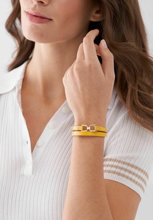 Femme ajustant ses cheveux bruns bouclés, portant une chemise blanche à manches courtes et un bracelet en cuir jaune à double tour avec fermoir en or.