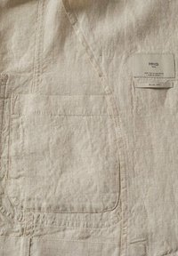 Beżowa tkanina o teksturze z kwadratową kieszenią i metką odzieżową po prawej stronie z napisem "MNG Man", rozmiar i "Slim Fit."