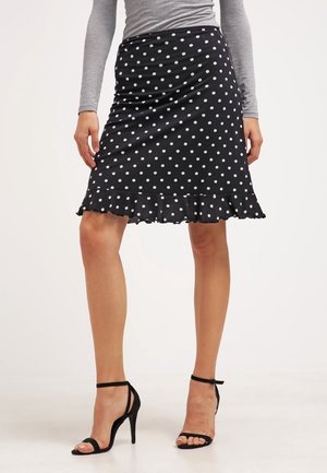 Kobieta ubrana w czarną spódnicę w kropki z falbaną na dole, szary długi rękaw i czarne sandały na obcasie z paskiem wokół kostki, stojąca.