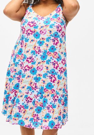 Femme portant une robe sans manches blanche ornée de motifs floraux vifs en bleu, violet et jaune sur un fond uni.