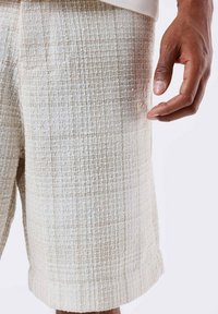 Shorts texturés beige avec un motif tissé, ourlet retroussé et poche latérale. Le matériau semble léger et décontracté. Une main tendue vers le tissu.