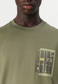 T-shirt vert olive à col rond, présentant un design de poche imprimé évoquant un conteneur maritime, avec du texte et des graphiques en noir et jaune.