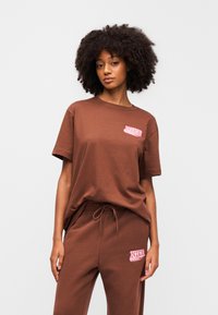 Καφέ βαμβακερό t-shirt με χαλαρή εφαρμογή, στρογγυλή λαιμόκοψη και κοντά μανίκια. Διαθέτει ροζ λογότυπο Nike στο στήθος. Συνδυάζεται με ταιριαστό παντελόνι φόρμας.