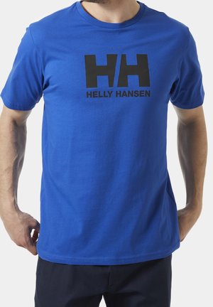 Mężczyzna w królewskim niebieskim t-shircie Helly Hansen z czarnym logo i napisem, stojący z rękami przy kieszeniach na białym tle.