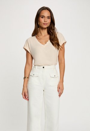 Femme aux longs cheveux bruns portant un haut beige à manches courtes avec encolure en V à bords festonnés et un pantalon blanc taille haute avec poches à rabat devant, debout devant un fond blanc.