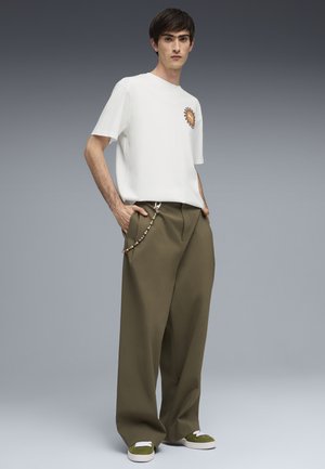 Jeune homme debout, les mains dans les poches, portant un T-shirt blanc avec un motif de soleil, un pantalon ample vert olive avec une chaîne, et des baskets vertes.