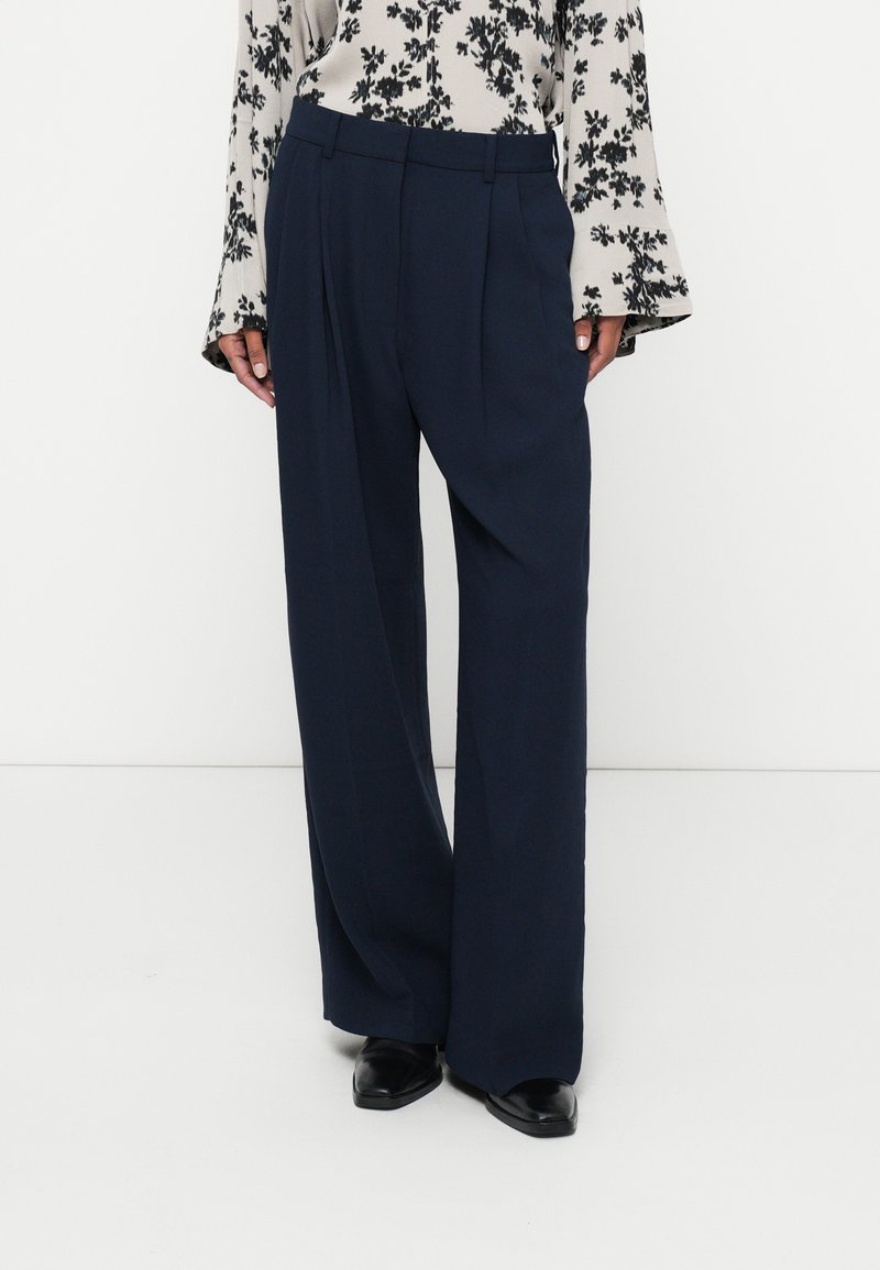 Pantalones de pierna ancha azul marino con una textura suave, frente plisado y cinturilla entallada. Combinados con una blusa floral de manga larga y zapatos negros.