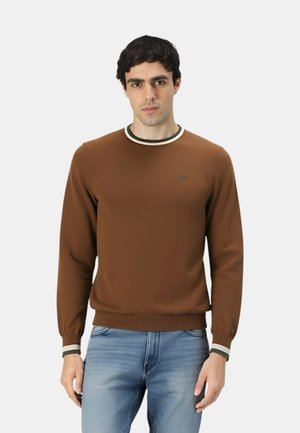 Uomo che indossa un maglione marrone a maniche lunghe con dettagli bianchi e neri al colletto e ai polsini, abbinato a jeans azzurri, in piedi su uno sfondo bianco.