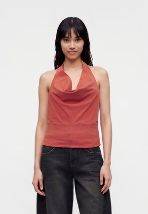 Ung kvinde med langt sort hår iført en ærmeløs rustfarvet haltertop og løse sorte jeans, står foran en ensfarvet hvid baggrund.
