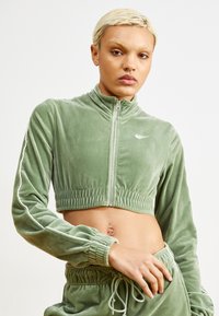 Chaqueta corta de terciopelo verde con cuello alto, cierre de media cremallera en el frente, cintura elástica y detalles de rayas blancas en las mangas.