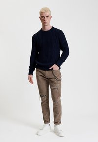Maglione blu navy con motivo a treccia, abbinato a pantaloni in corduroy beige con strappi e dettagli in catena. Le scarpe da ginnastica bianche completano il look.