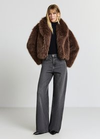 Giacca corta in pelliccia sintetica marrone con ampio colletto, abbinata a jeans grigio scuro a vita alta. Modella in piedi su uno sfondo neutro.