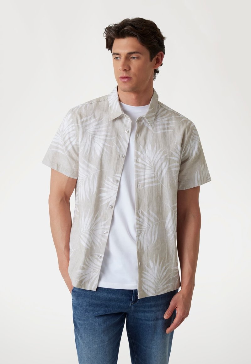 Camisa de manga corta en un beige claro con sutiles estampados de hojas blancas, cierre de botones y un corte relajado, combinada con una camiseta blanca debajo.
