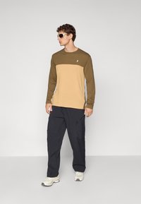 Skjorte med lange ermer har en brun øverste del og en lys tan nederste del, kombinert med mørke cargo-bukser og joggesko. Stylet med solbriller.