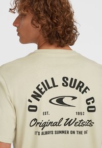 Lichtbeige t-shirt met zwarte tekst en logo op de achterkant, met de nadruk op "O'Neill Surf Co" en "Original Wetsuits." Zachte katoen textuur.