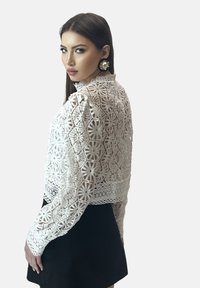 Giacca corta in pizzo bianco con motivo floreale intricato, colletto alto e maniche lunghe. Abbinata a una gonna nera, che mette in mostra un design dettagliato.