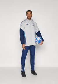 Grå og marineblå treningsdress med hvite detaljer, jakke med glidelås og tapered bukser. Holder en flerfarget fotball.