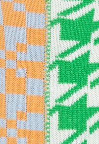 Tessuto a maglia diviso in due sezioni: un lato presenta motivi a scacchi arancioni e blu, mentre l'altro mostra forme geometriche di foglie verdi su sfondo bianco.