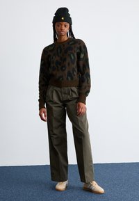 Lepelprint gebreide trui in bruin en groen, gecombineerd met olijfgroene broek en beige sneakers met witte strepen; zwarte beanie.