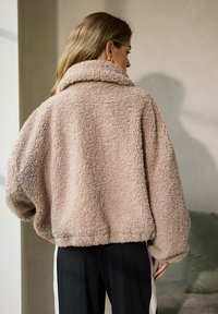 Softe taupe shearling jakke med høj krave, oversized ærmer og elastiske cuffs. Bæret sammen med sorte bukser med hvide sidelister.