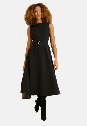 Žena s kudrnatými vlasy, která má na sobě černé midi šaty bez rukávů s páskem a kapesami, doplněné černými botami na podpatku, stojící na bílém pozadí.