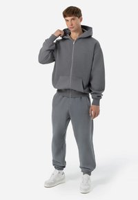 Felpa grigia con zip, dotata di tasche frontali e pantaloni da tuta grigi abbinati. Il tessuto sembra morbido, con polsini e vita elasticizzati.