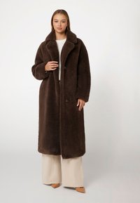 Abrigo de lana marrón largo con cuello de muesca, cierre de botones y corte holgado. Combinado con pantalones de pierna ancha de color claro y tacones de punta.