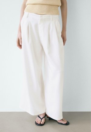 Persona che indossa pantaloni bianchi larghi a vita alta, top beige senza maniche in maglia e sandali neri con punta quadrata, in piedi contro una parete chiara.