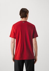 Röd bomullströja med korta ärmar. Har en rund hals och en liten grön Lacoste-logga på baksidan. Slät textur och avslappnad passform.