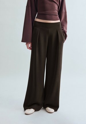 Osoba nosiaca široké čierne nohavice, hnedý crop top s dlhým rukávom a biele topánky, stojaca pred jednoliatym svetlým pozadím.