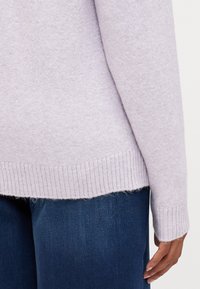 Suéter de malha em lavanda clara com punhos e bainha canelados. O tecido parece suave e ligeiramente texturizado, combinado com calças de ganga escuras.