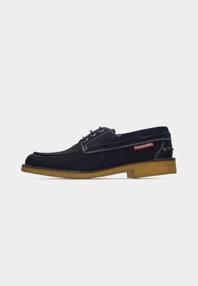 Chaussure bateau en daim bleu marine avec semelle beige, coutures blanches, trois œillets pour les lacets et étiquette rouge DSQUARED2 sur le côté.