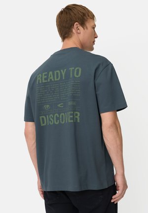 Mand iført en mørkeblå t-shirt med grøn tekst "READY TO DISCOVER" trykt på bagsiden, vendt væk fra kameraet.