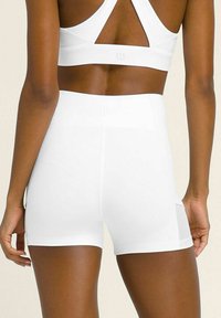 Donna che indossa pantaloni sportivi bianchi a vita alta e un reggiseno sportivo bianco, vista di spalle su uno sfondo neutro.