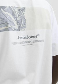 Bílá bavlněná trička s šedým grafickým designem na hrudi a značkou "Jack & Jones" vytištěnou pod ním černou barvou.